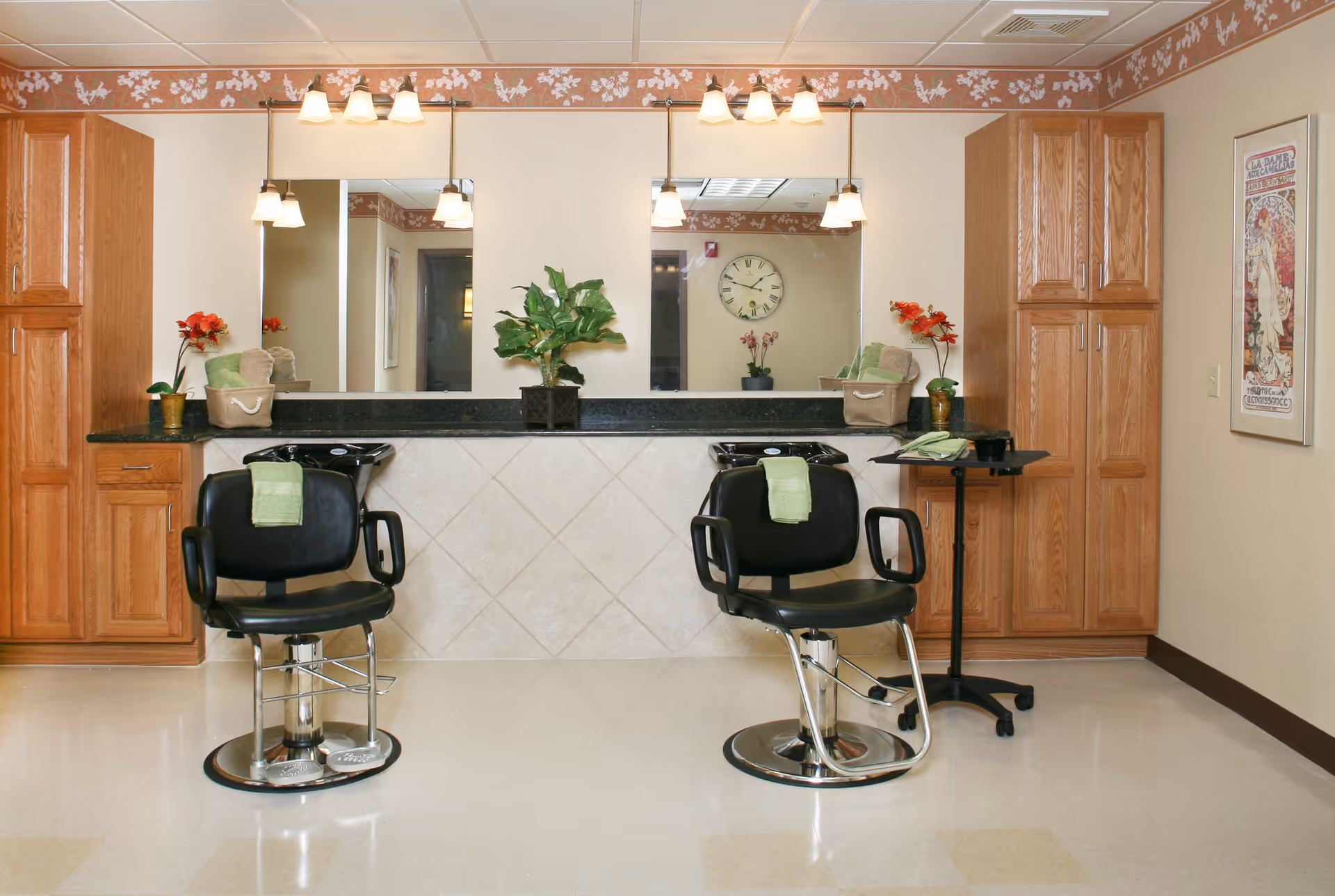 Two black salon chairs in front of mirrored styling stations with wooden cabinets, plants, and overhead lights in a salon area.