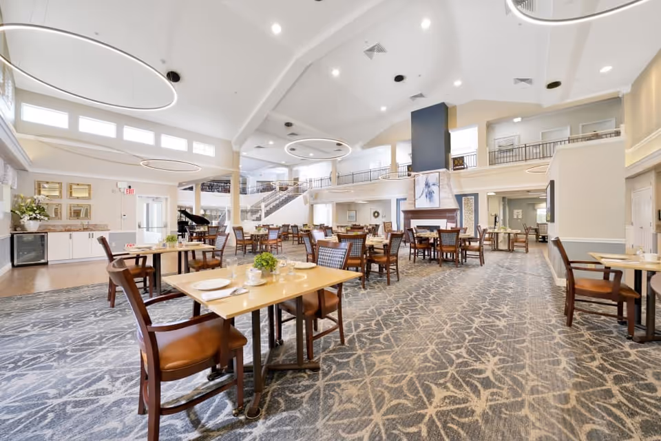 Spacious dining room with multiple wooden tables and chairs, high ceilings, circular light fixtures, and a central fireplace.