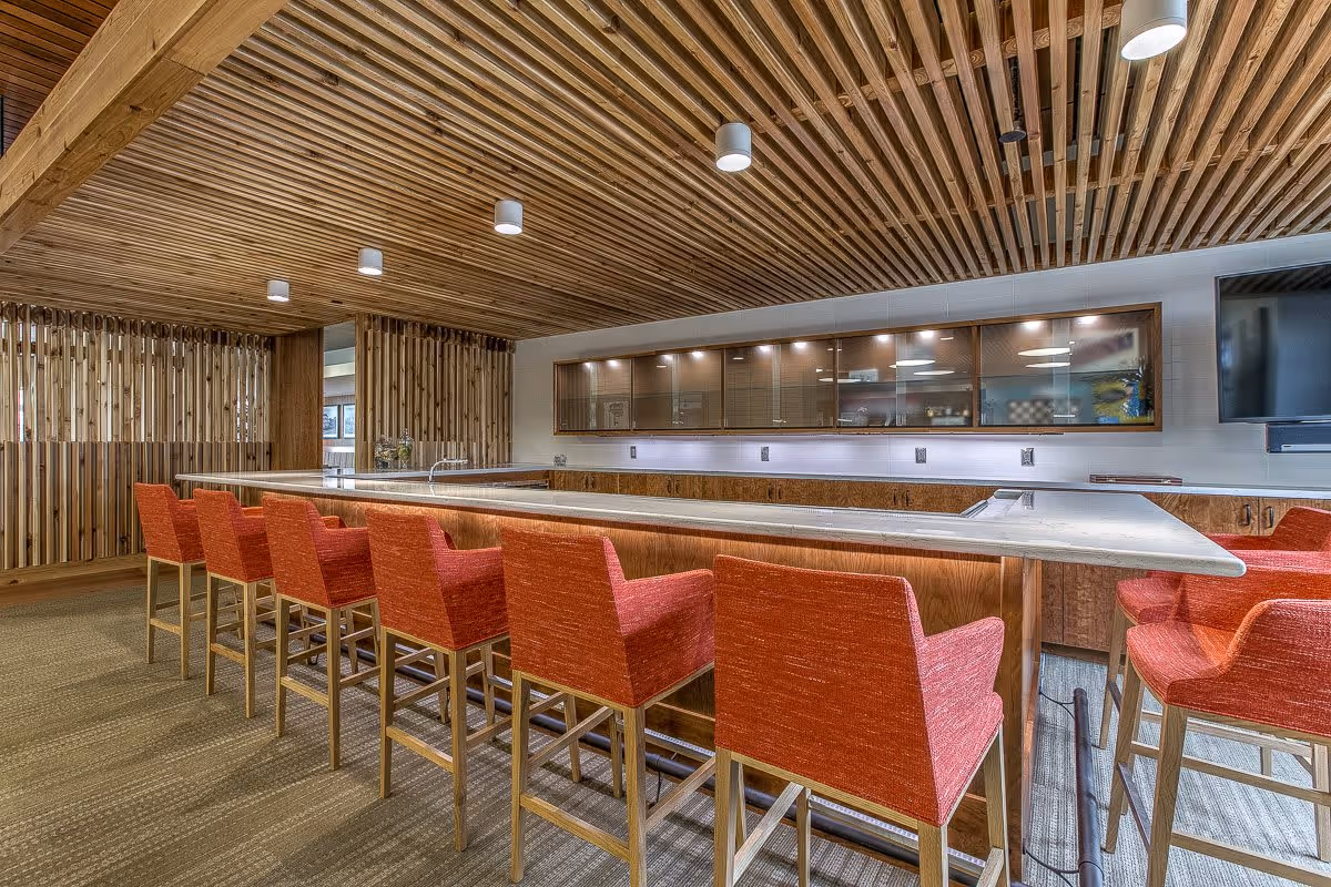 Interior communal bar area with a long countertop and red upholstered bar stools under a wooden slatted ceiling.