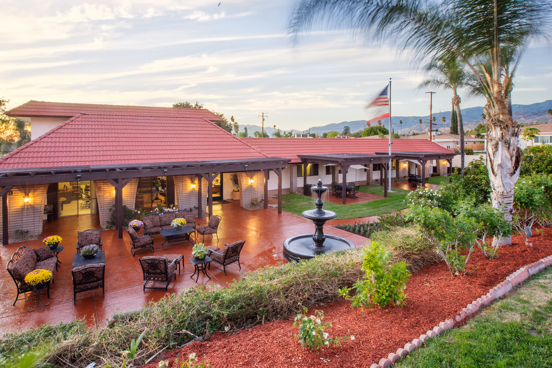 Outdoor patio area of Highland Palms Healthcare Center featuring a red-tiled roof building with covered seating areas, multiple chairs and tables with flower pots, a central black fountain, landscaped garden beds with shrubs and flowers, palm trees, and an American flag on a flagpole. Mountains and a partly cloudy sky are visible in the background.
