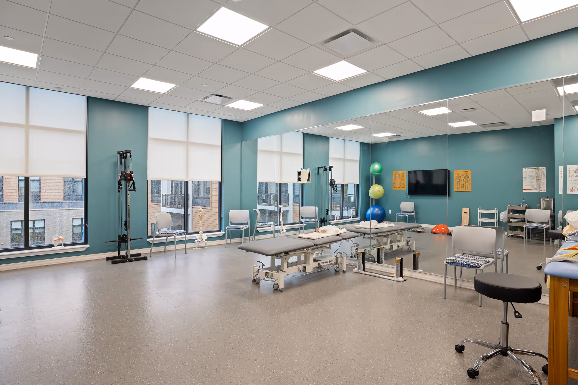 A spacious physical therapy or rehabilitation room with teal walls, large windows with white blinds, a large wall mirror, therapy tables with pillows, exercise balls stacked in a corner, chairs, and various therapy equipment.