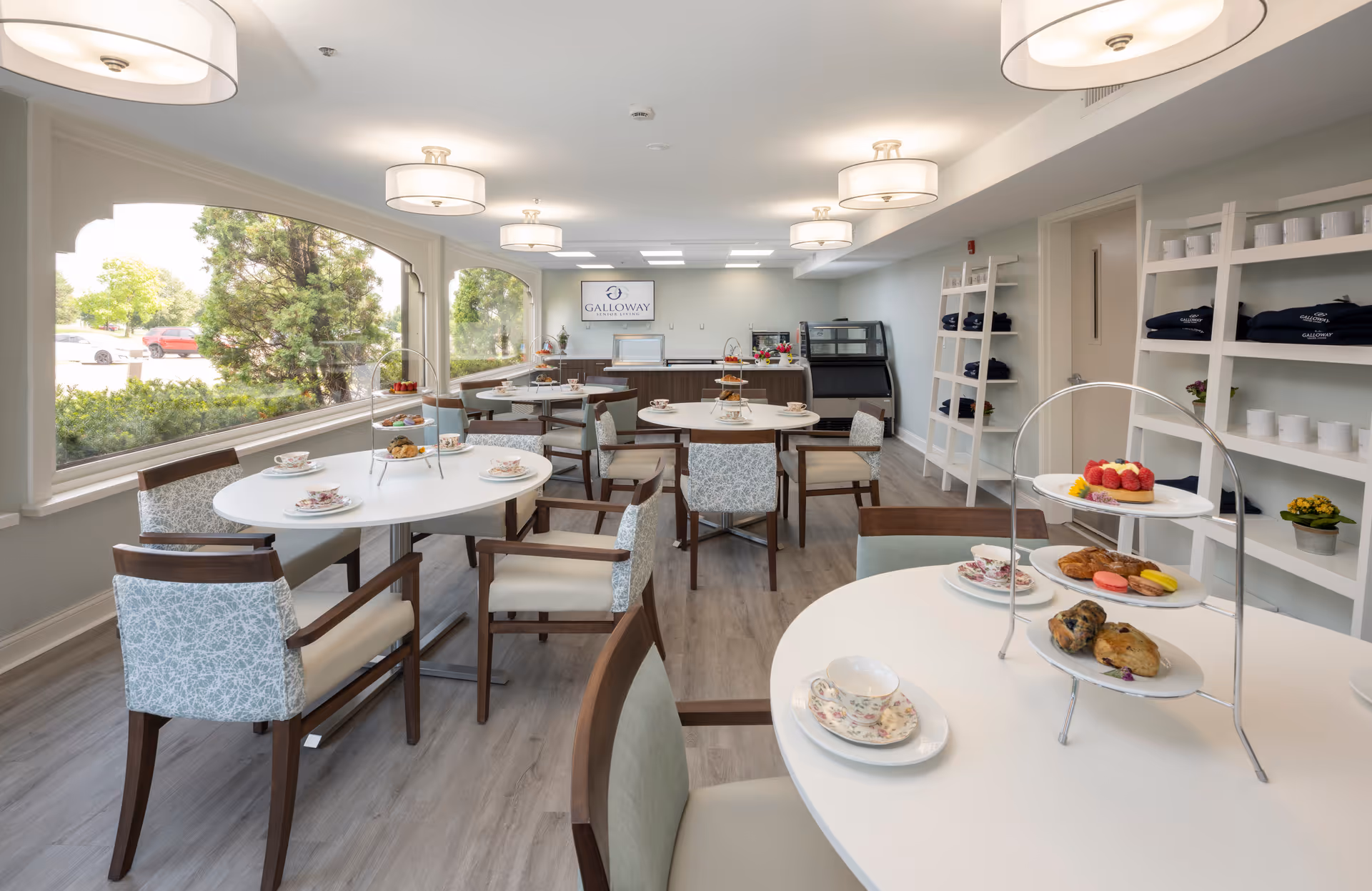 A bright and airy dining room in a senior living facility with round tables set with floral teacups and tiered trays of pastries and desserts. The room features large windows with views of greenery outside, modern light fixtures on the ceiling, wooden chairs with cushioned seats, and white shelving units holding folded blankets and mugs. A sign on the back wall reads 'Galloway'.