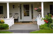 Front porch of a white house with a central door, two chairs, a hanging flower basket, and a bicycle leaning against the railing.