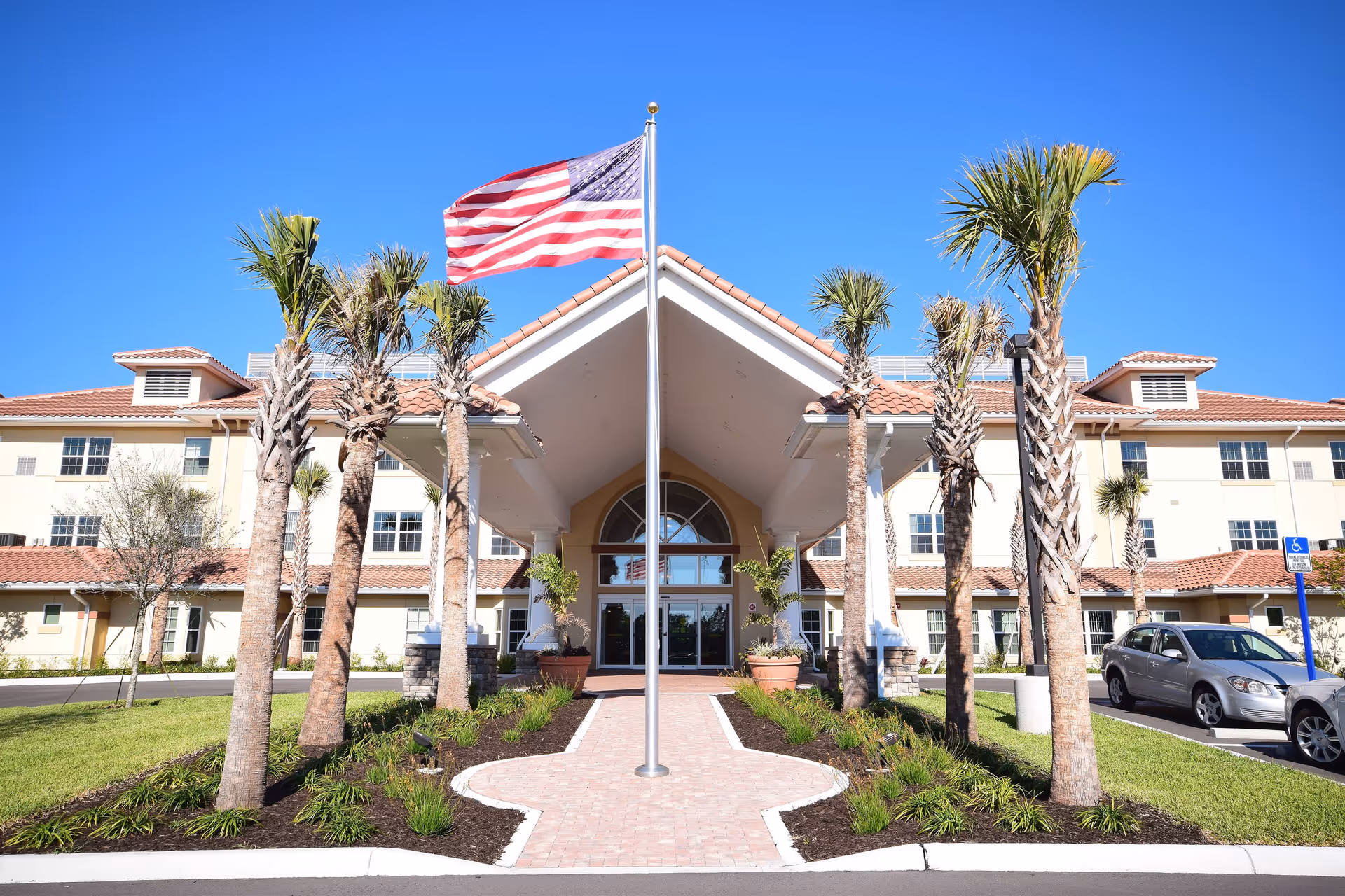 Front exterior view of Stone River Retirement Community building with a covered entrance, palm trees lining the walkway, an American flag on a flagpole in the center, and parked cars on the right side under a clear blue sky.