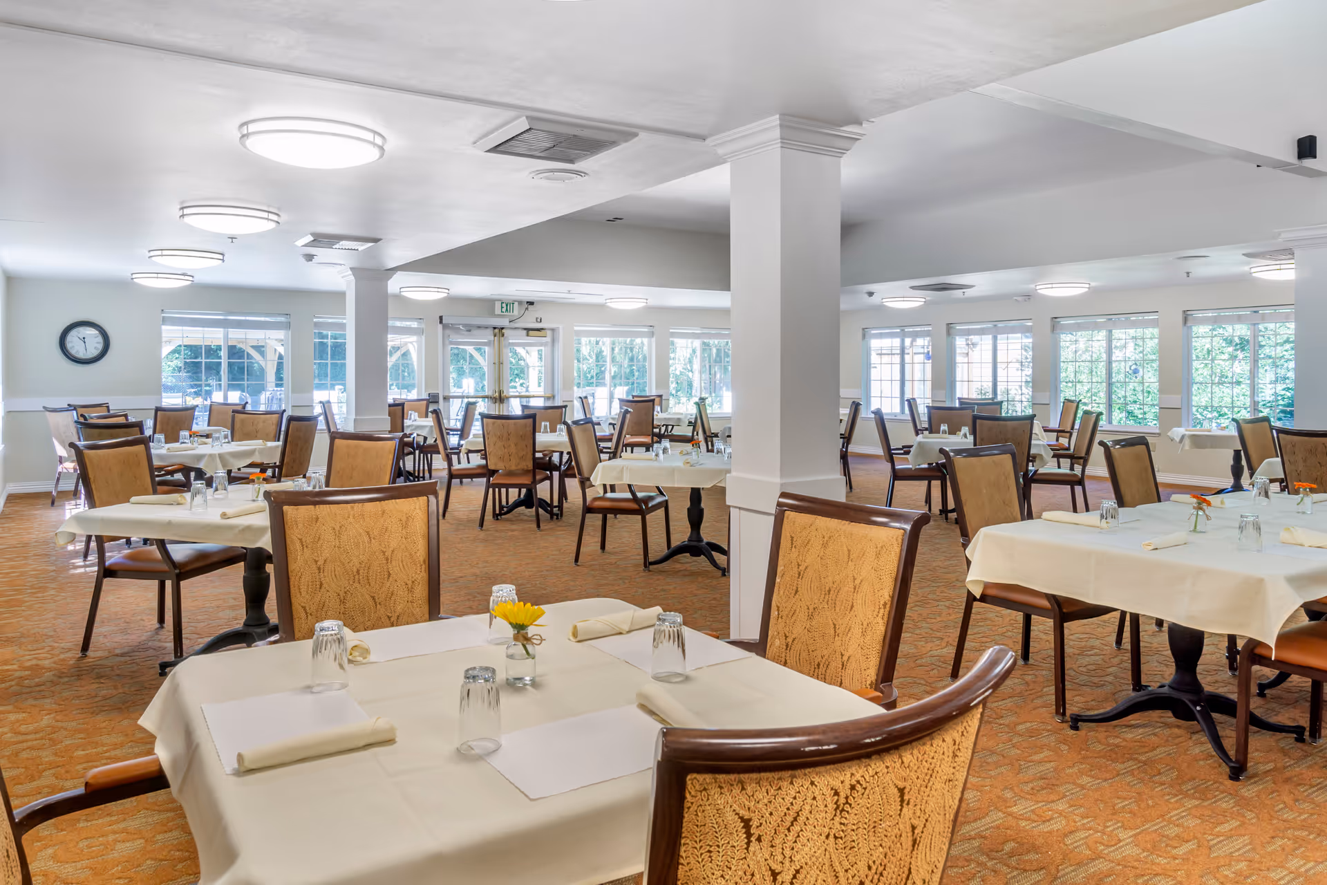 Bright, spacious dining room with multiple tables set with white tablecloths and chairs and large windows letting in natural light.