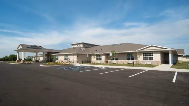 Single-story memory care building with a covered entrance and empty parking lot under a clear blue sky.