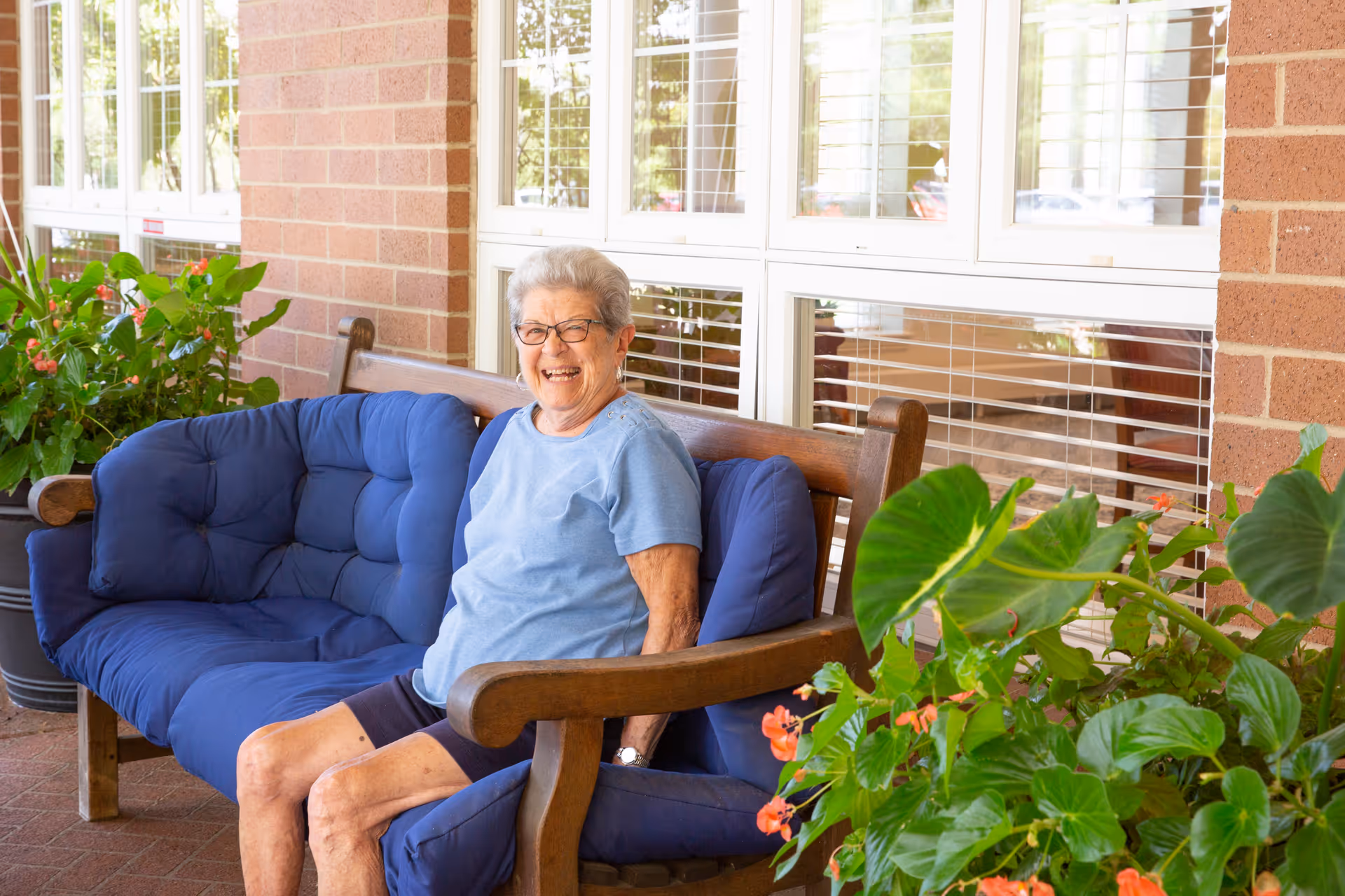 An elderly woman wearing glasses and a light blue shirt sits smiling on a cushioned wooden bench outside a building with large windows. There are green plants with orange flowers around her, and the setting appears to be a patio or outdoor seating area.
