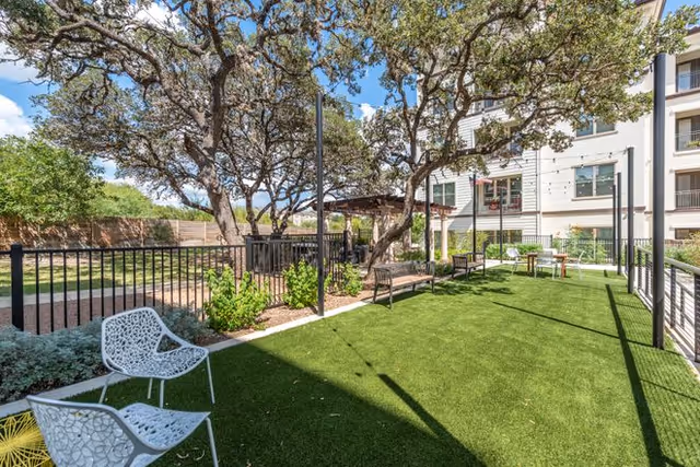 Outdoor area at Overture Stone Oak featuring artificial grass, several benches, white metal chairs, trees providing shade, and a multi-story building in the background under a blue sky with some clouds.