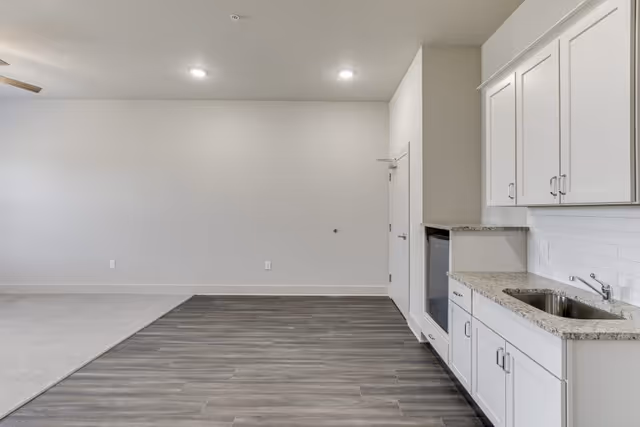 Interior view of a room with a kitchenette on the right side featuring white cabinets, a sink, and granite countertops. The floor transitions from wood-style flooring near the kitchenette to carpeted flooring on the left. The walls are painted light gray, and there are recessed ceiling lights and a ceiling fan partially visible on the left.