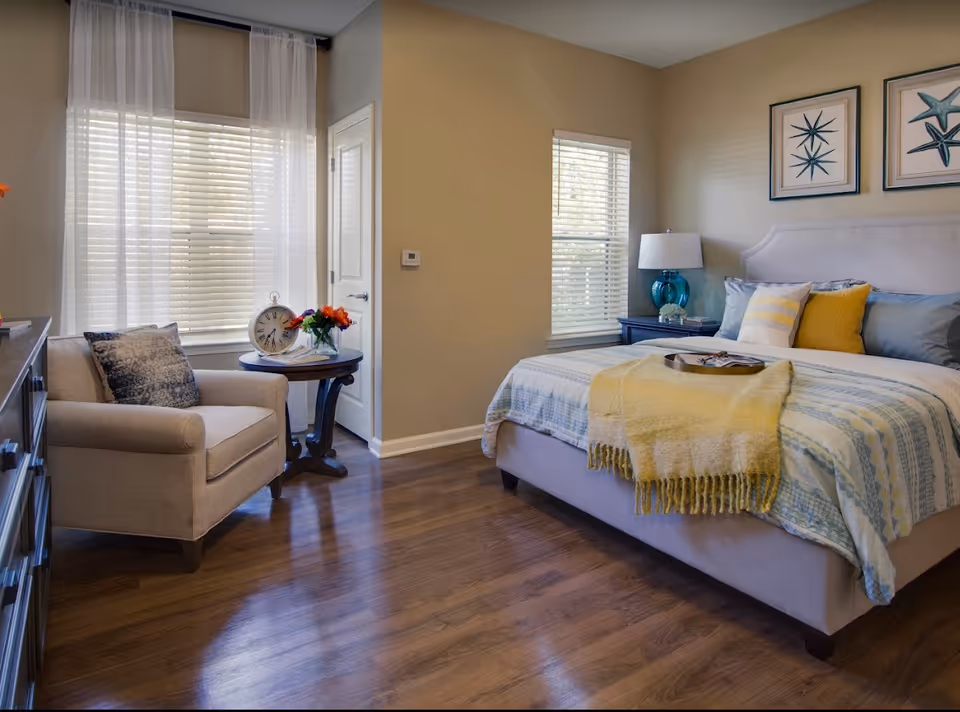 A cozy bedroom with a large bed covered in a light blue and yellow patterned blanket and multiple pillows. A yellow throw blanket is draped over the foot of the bed with a tray on top. Next to the bed is a dark blue nightstand with a blue glass lamp and some books. On the wall above the bed are two framed starfish artworks. To the left, there is a beige armchair with a patterned pillow and a round wooden side table holding a large clock and a vase of flowers. The room has wooden flooring and two windows with blinds and sheer white curtains.