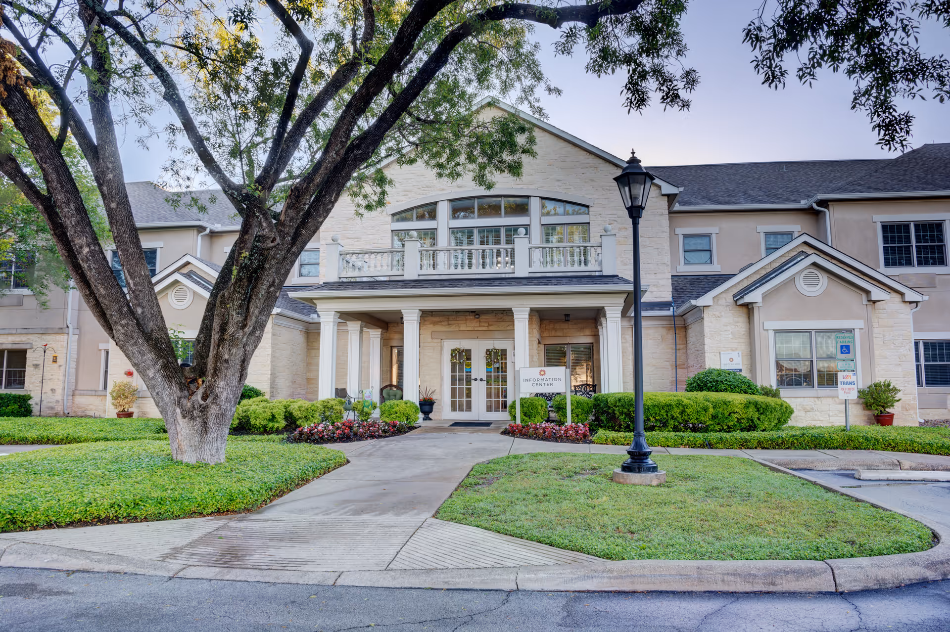 Front exterior view of Gemstone Senior Living at Castle Hills showing a large tree in the foreground, a paved walkway leading to the entrance with white columns, manicured bushes and flower beds, and a lamppost near the sidewalk.