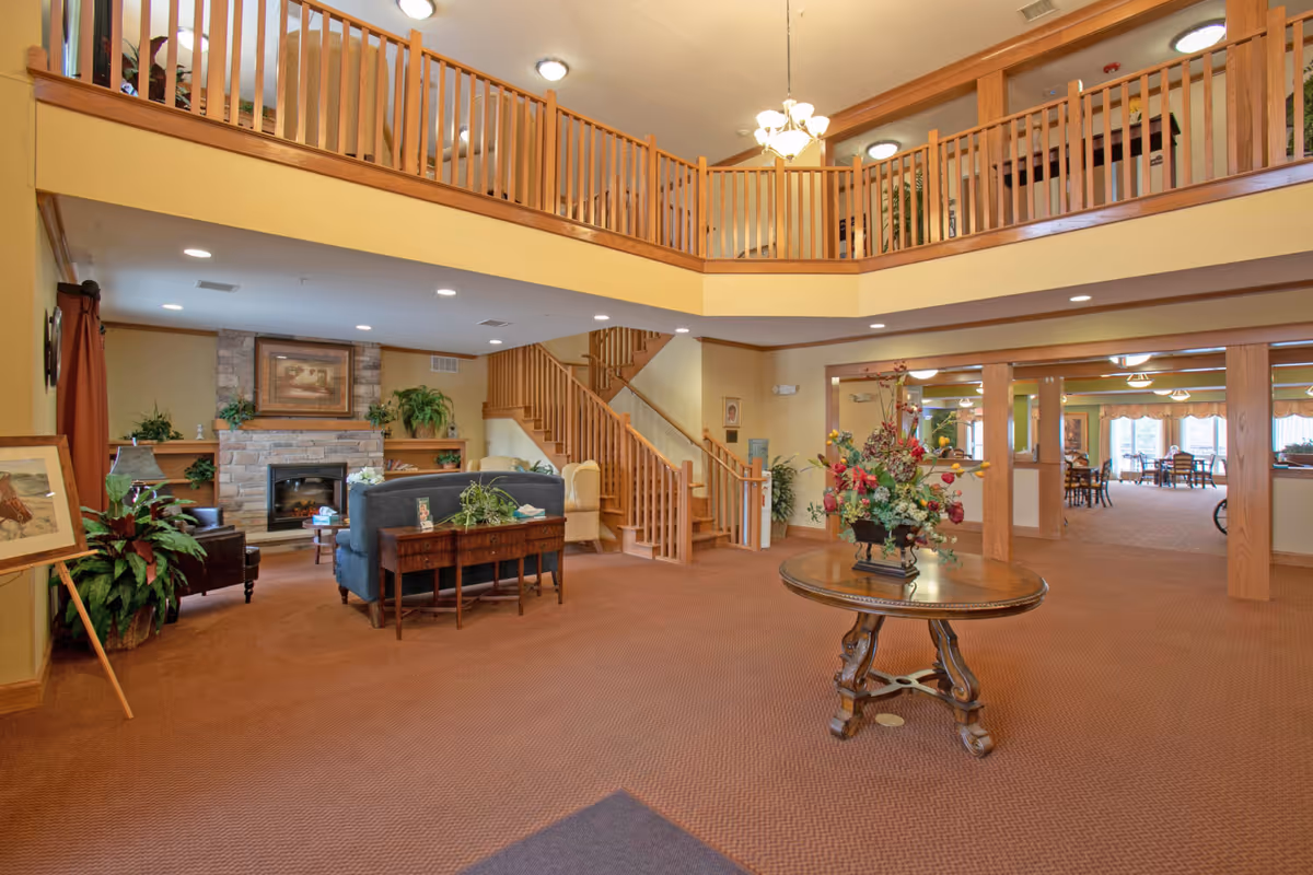 Spacious two-story lobby/lounge with seating around a fireplace, a central round table topped with a floral arrangement, and a wooden staircase and balcony.