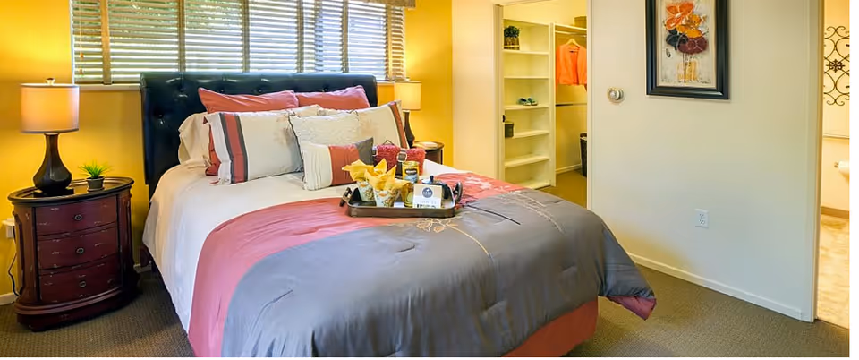 A cozy bedroom with a neatly made bed featuring a gray and pink comforter and multiple pillows. There are two bedside tables with lamps on each side of the bed. Behind the bed is a window with blinds. To the right, there is an open walk-in closet with shelves and hanging clothes, and a doorway leading to a bathroom.