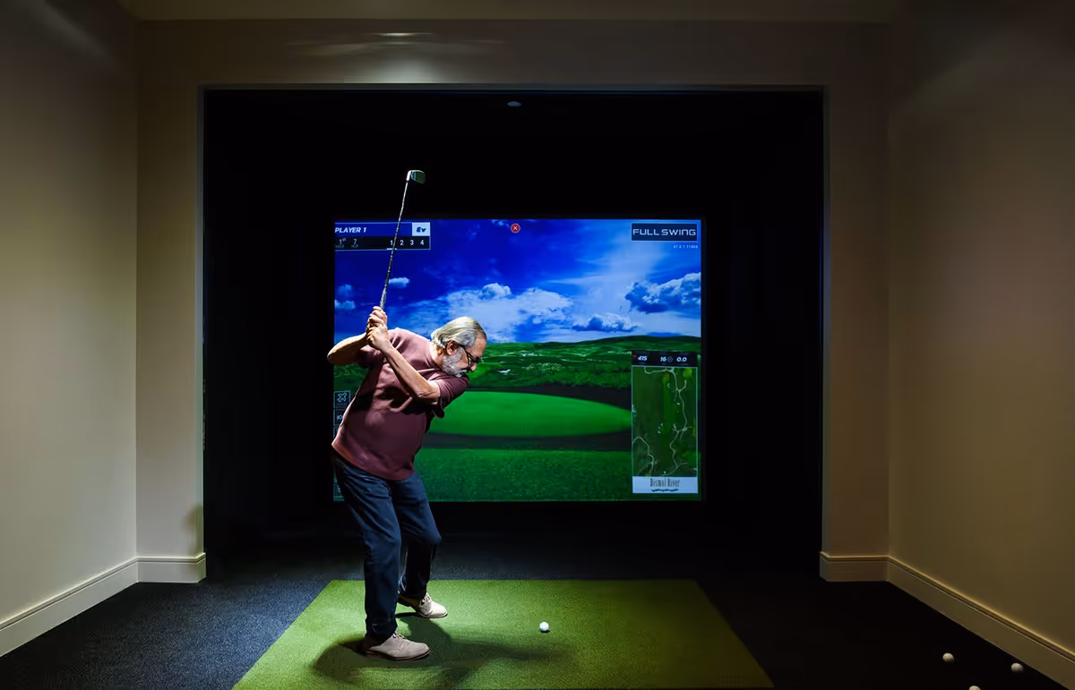 An elderly man with gray hair and a beard is playing indoor golf using a golf simulator. He is mid-swing on a green mat in a dimly lit room with a large screen displaying a golf course in front of him.
