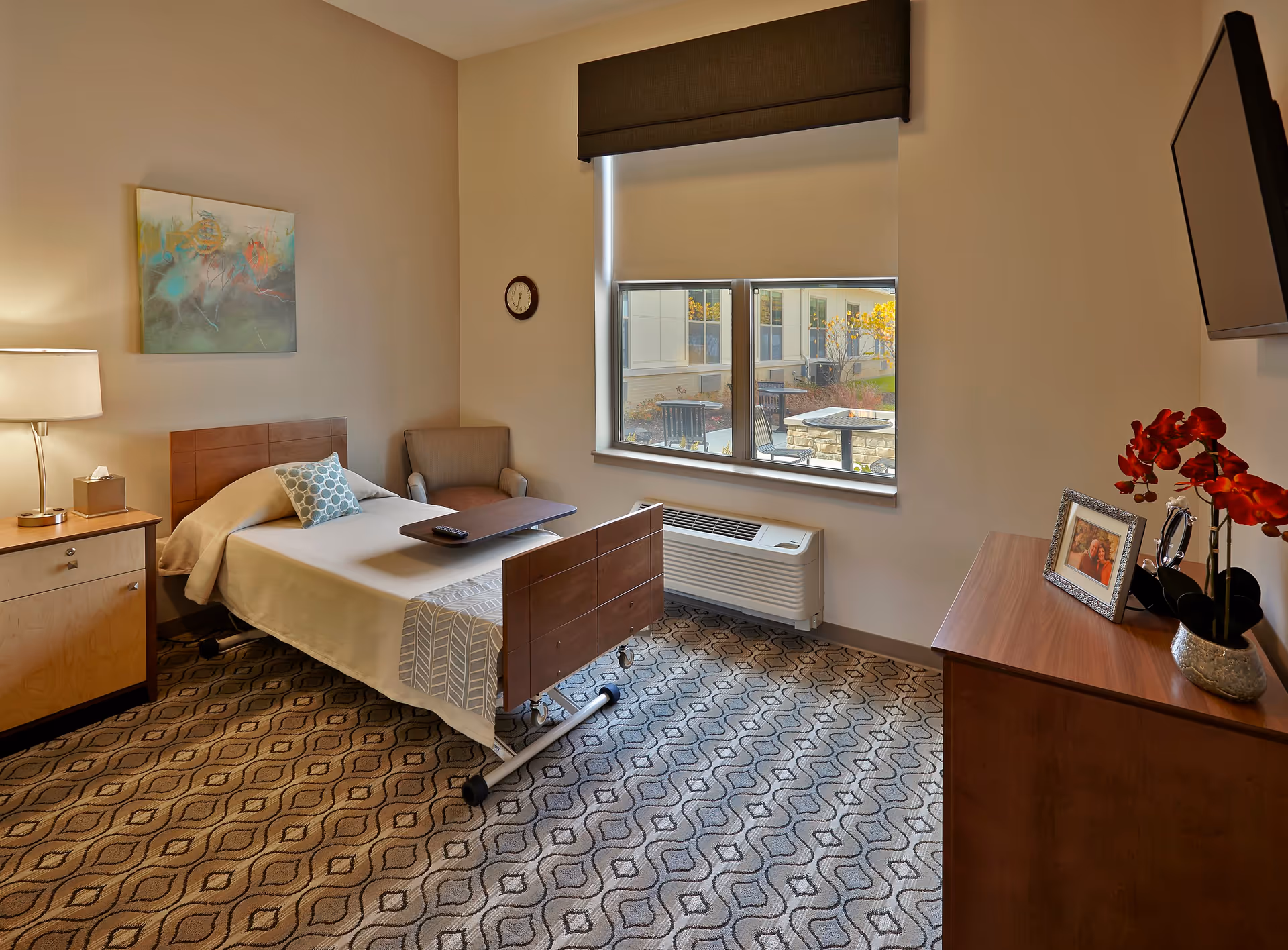 Furnished senior living bedroom with a single bed, nightstand, armchair, dresser, wall-mounted TV and a window overlooking an outdoor patio.