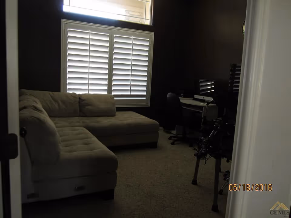 A dimly lit room with a beige sectional sofa on the left side, a window with white plantation shutters in the center, and a desk with a computer and office chair on the right side. The room has carpeted flooring and dark walls.