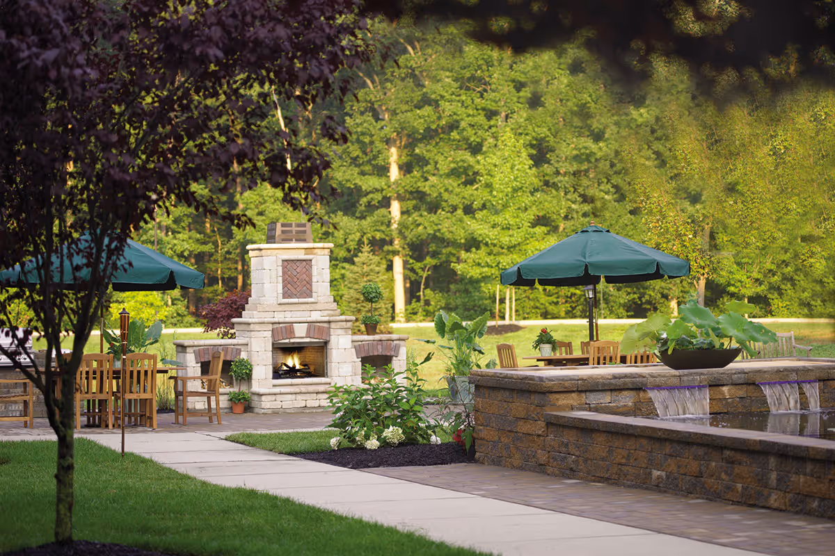 Outdoor patio area with a stone fireplace, wooden tables and chairs under green umbrellas, surrounded by lush green trees and plants, with a stone water feature in the foreground.
