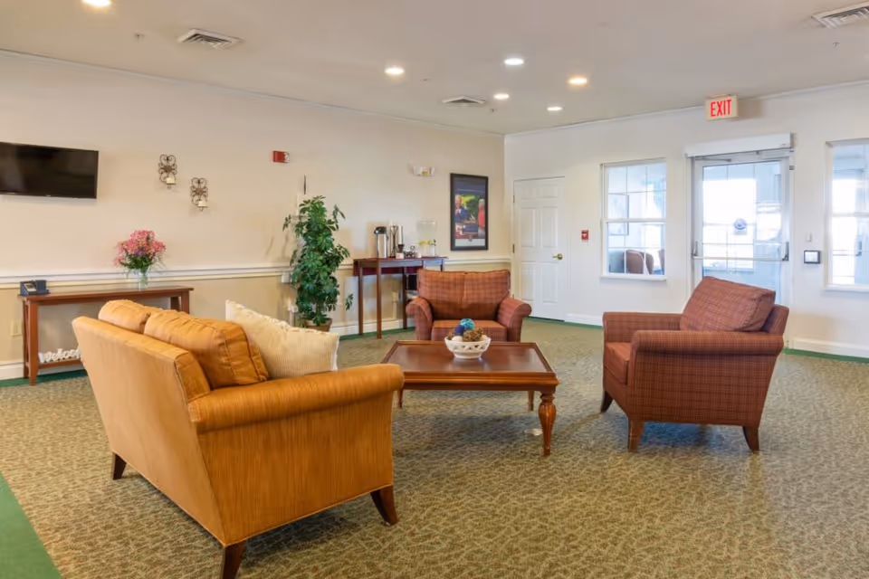 Seating area with sofas and armchairs arranged around a coffee table in a well-lit common room with a TV and exit doors.