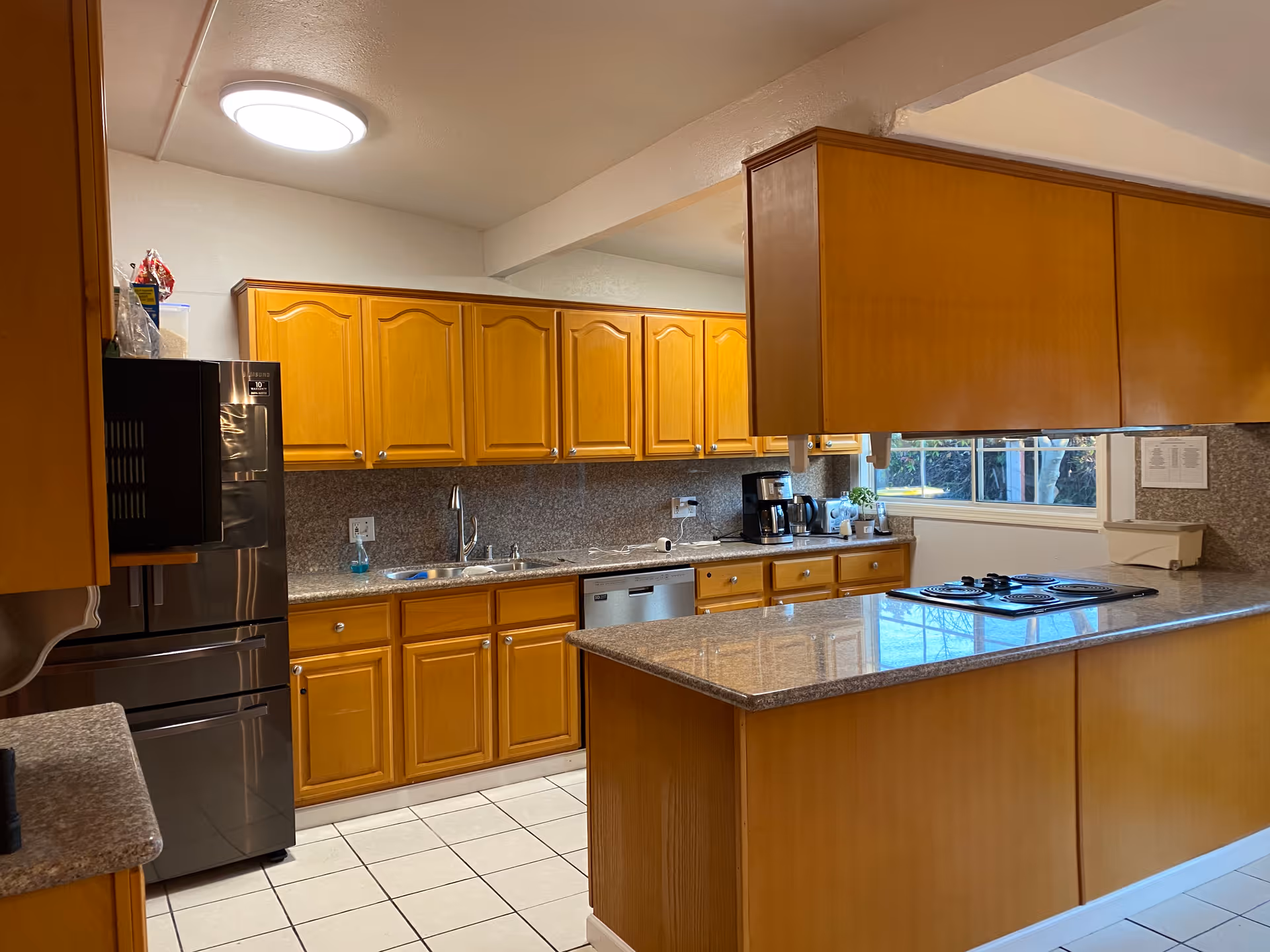 A spacious kitchen with wooden cabinets, granite countertops, a stainless steel refrigerator, a dishwasher, a coffee maker, and a stovetop on the kitchen island. There is a window above the counter letting in natural light.