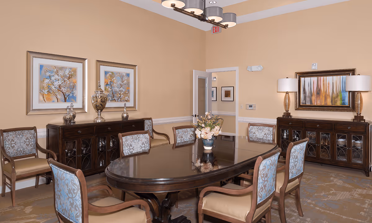 A formal dining room with a large dark wood oval table surrounded by eight upholstered chairs with patterned backs. Two dark wood sideboards with glass doors are against the walls, each decorated with lamps, vases, and framed artwork. The walls are painted beige, and there is a modern chandelier hanging from the ceiling. A door is open, showing a glimpse of an adjacent room with framed pictures on the wall.