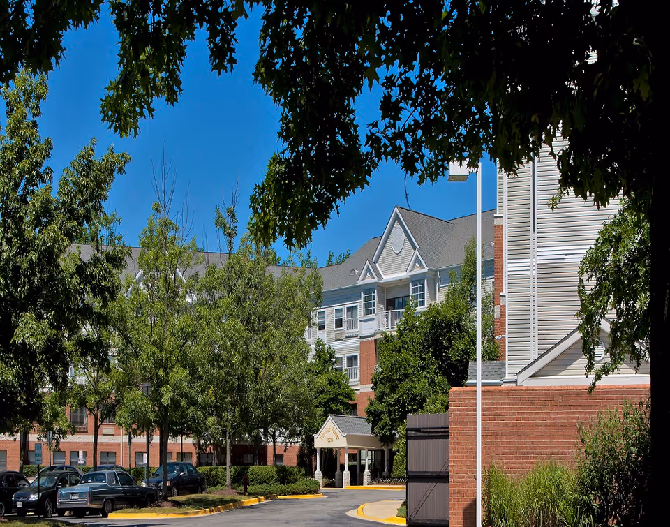 Exterior view of a multi-story senior living facility named Pin Oak Village, with a driveway entrance, several parked cars, and trees lining the area under a clear blue sky.