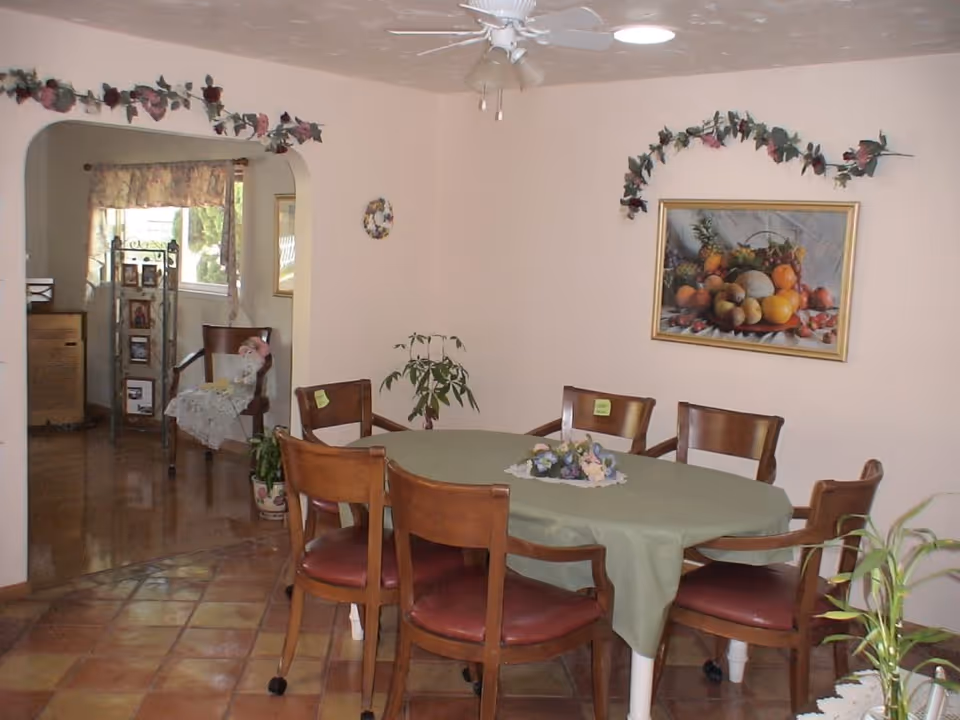 A dining room with a round table covered with a green tablecloth and six wooden chairs with red cushions. The walls are light-colored and decorated with artificial flower garlands and a framed painting of fruit. There is a ceiling fan above the table. In the background, an archway leads to another room with a chair, a small table, and a window with floral curtains.