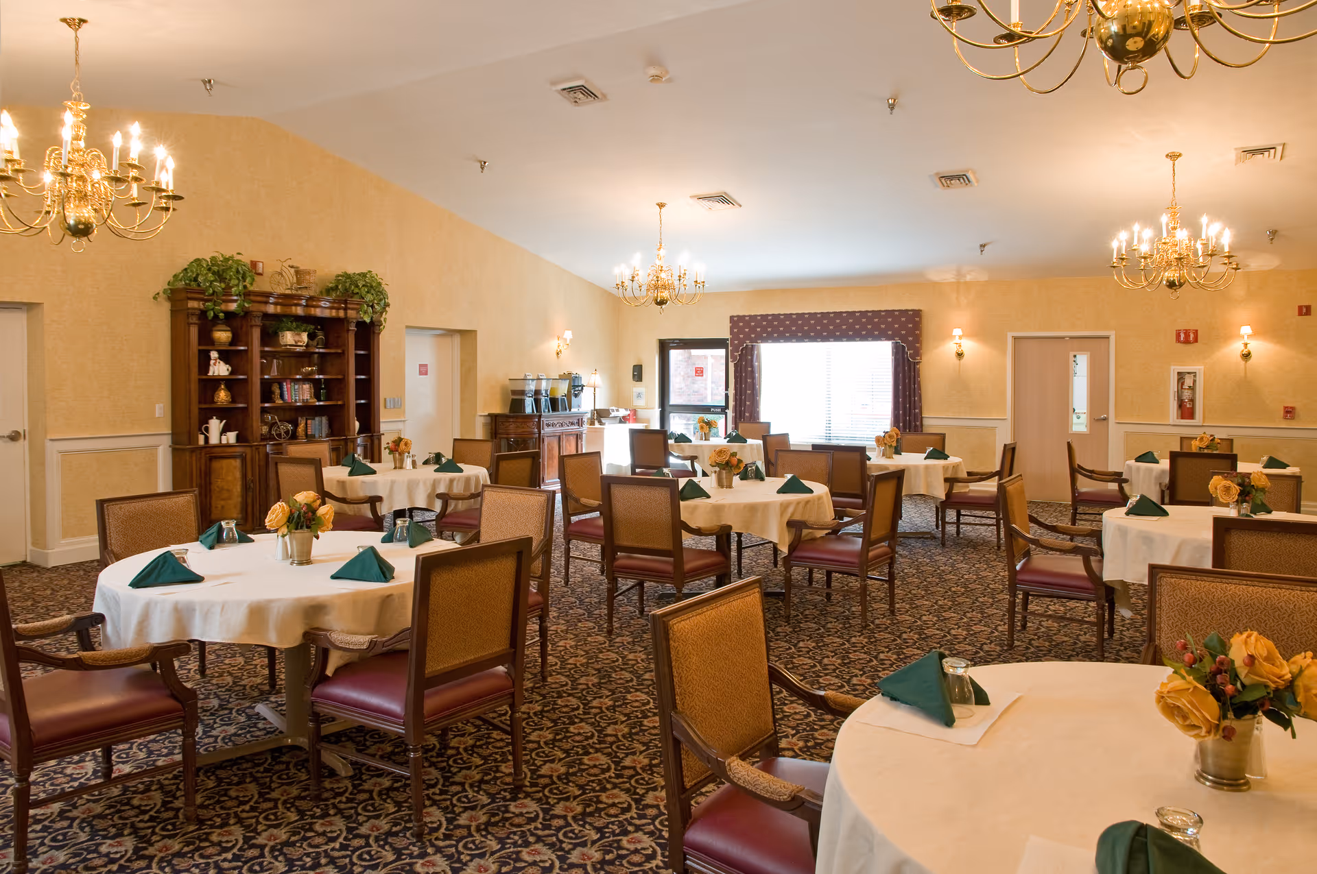 Dining room with round tables set with white tablecloths, green napkins, floral centerpieces, chandeliers, and upholstered chairs.