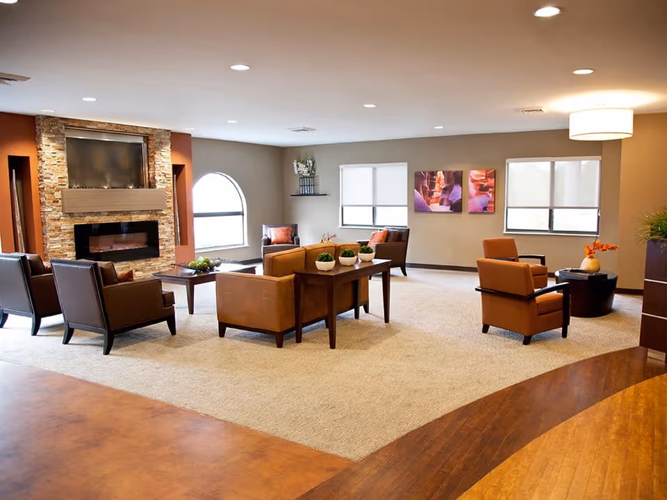 A spacious and well-lit living room area in a senior living facility featuring a stone fireplace, multiple comfortable armchairs and sofas arranged around coffee tables, large windows with blinds, decorative plants, and warm-toned walls and flooring.