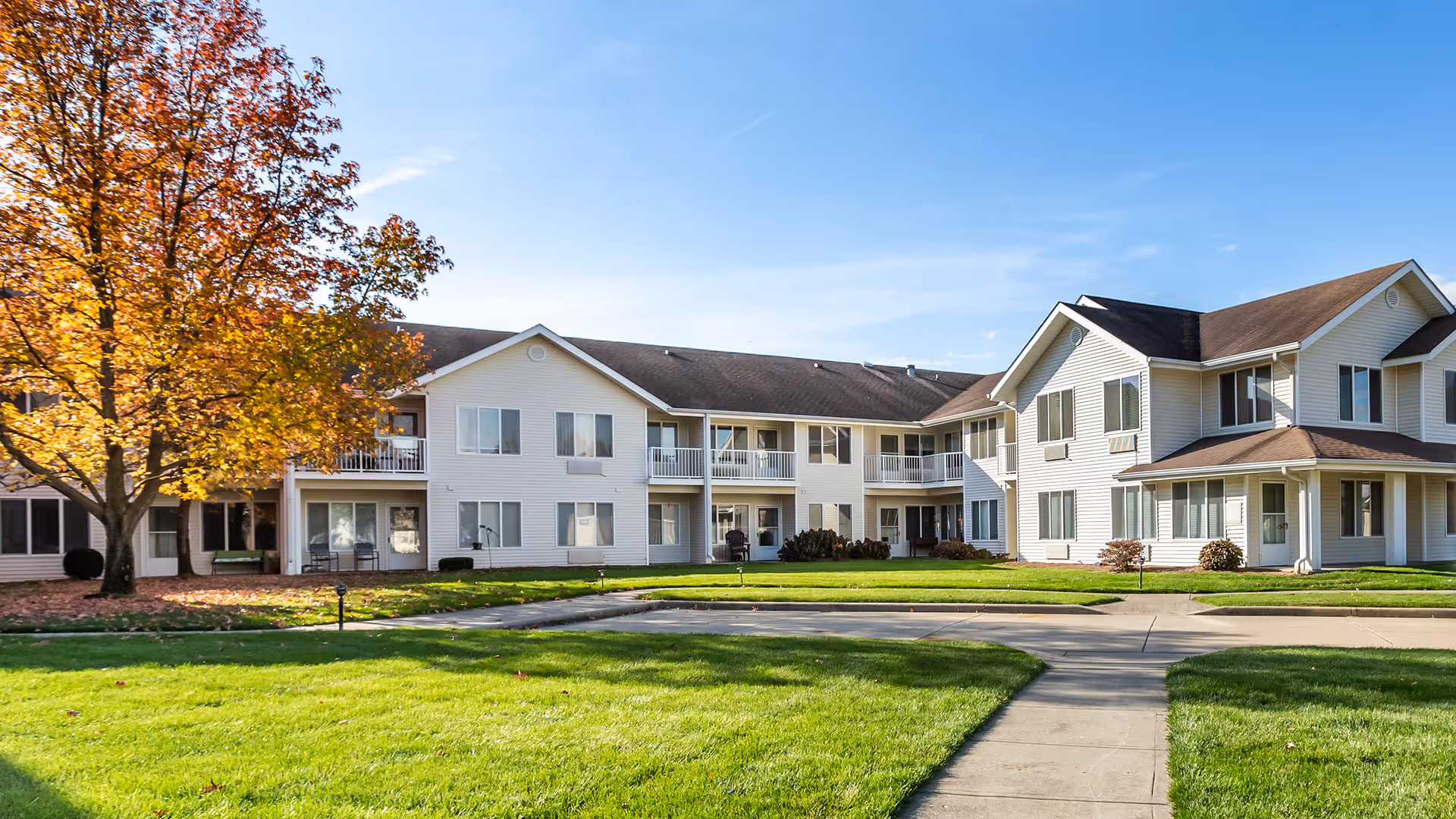 Exterior view of a two-story senior living facility building with white siding, multiple windows, and balconies. There is a well-maintained green lawn and a large tree with autumn-colored leaves in front of the building under a clear blue sky.