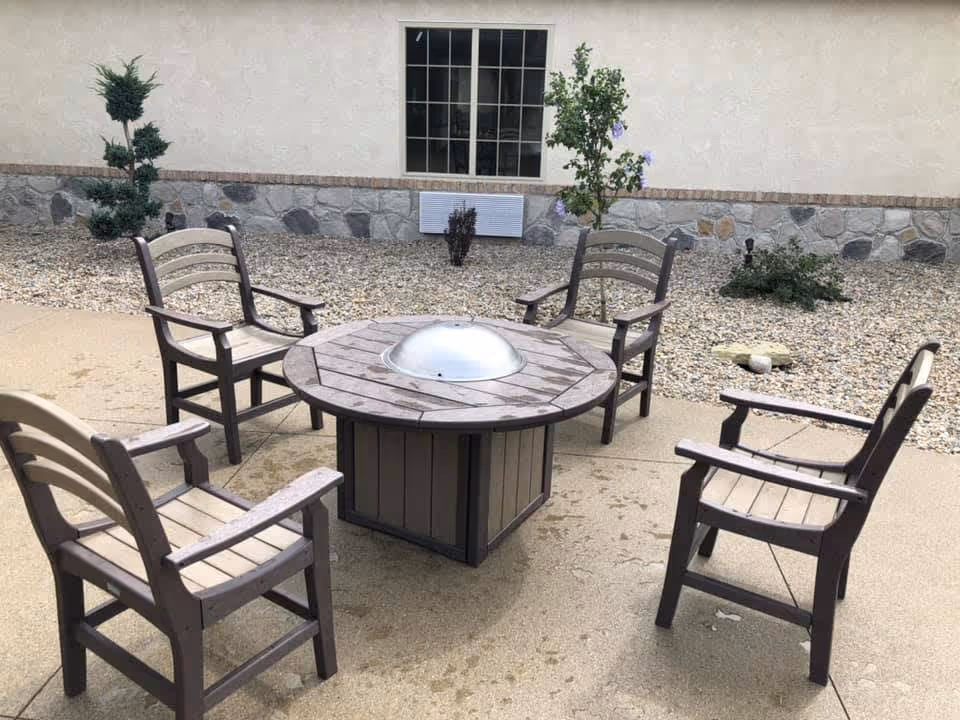 A round firepit table surrounded by five wooden armchairs on a paved outdoor patio in front of a building wall with gravel landscaping.