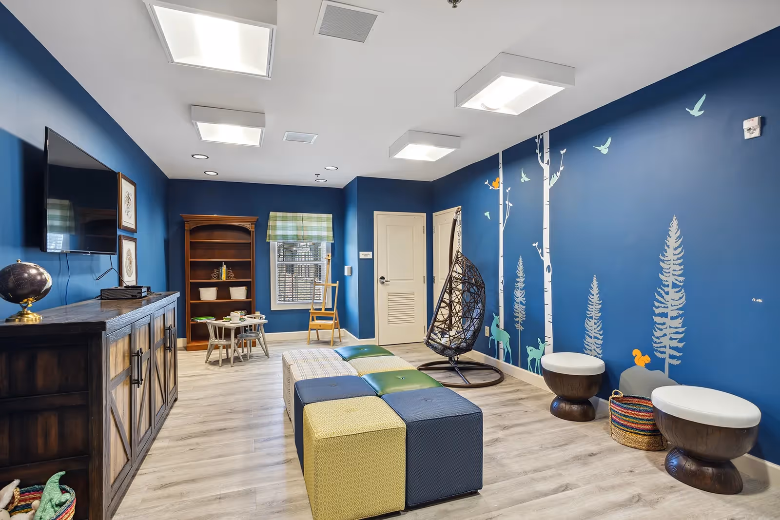 A bright and colorful common room with blue walls decorated with tree and bird decals. The room features a hanging wicker chair, two small round stools with white cushions, a multicolored cushioned bench, a wooden cabinet with a globe on top, a wall-mounted TV, a small table with chairs, and a window with a green plaid valance.