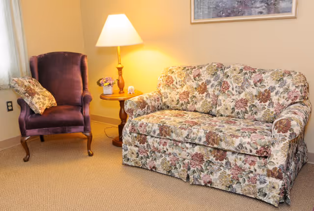A cozy living room corner featuring a floral-patterned sofa and a purple upholstered armchair with a matching floral pillow. Between them is a wooden side table with a lit table lamp and a small flower arrangement. The walls are painted a soft beige color, and a framed picture hangs above the sofa.