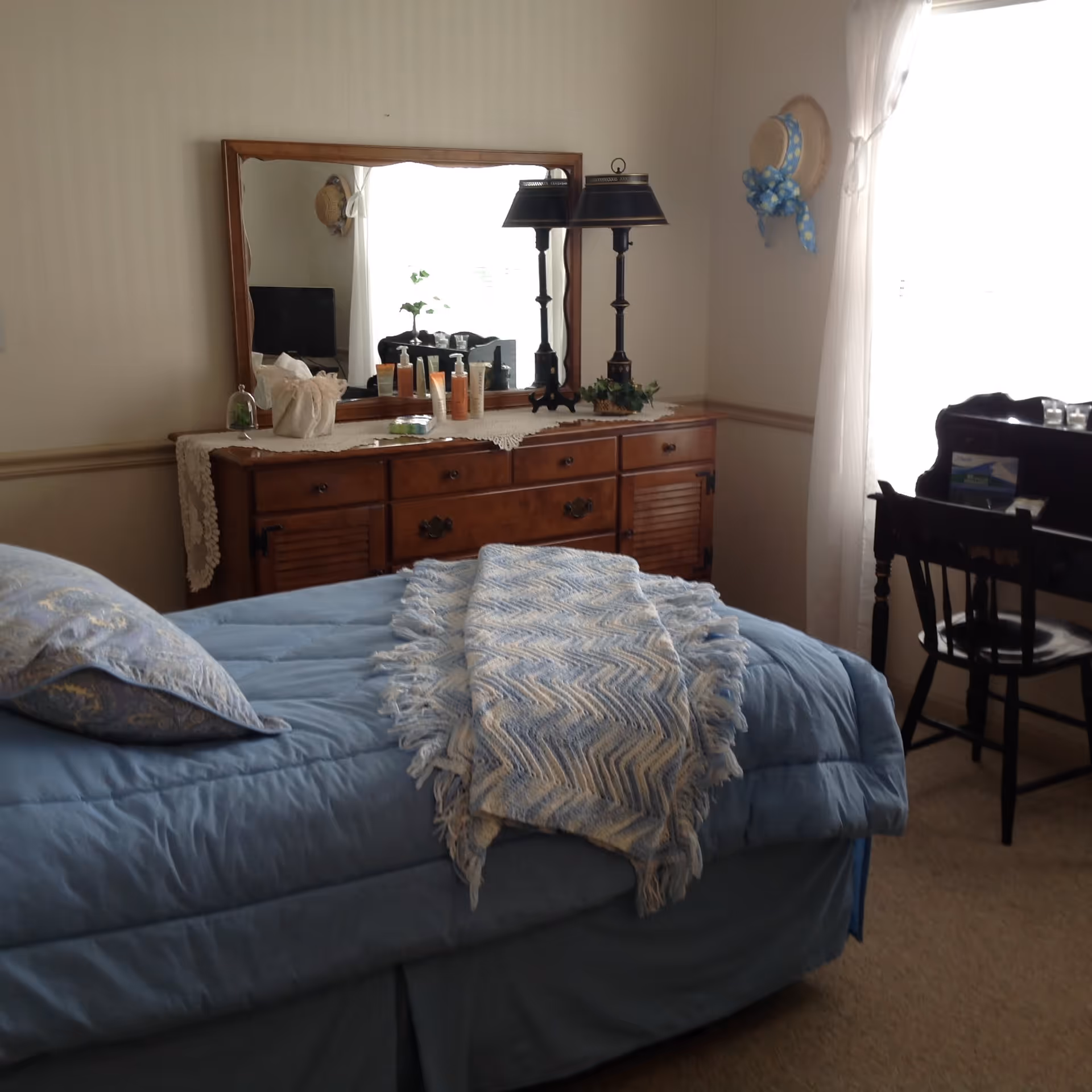 A cozy bedroom with a blue bedspread and a patterned throw blanket on the bed. There is a wooden dresser with a large mirror above it, decorated with a lace runner, a lamp, and various small items. A window with sheer white curtains lets in natural light, and a black chair is positioned next to a small desk or vanity. A decorative hat with a blue ribbon hangs on the wall.