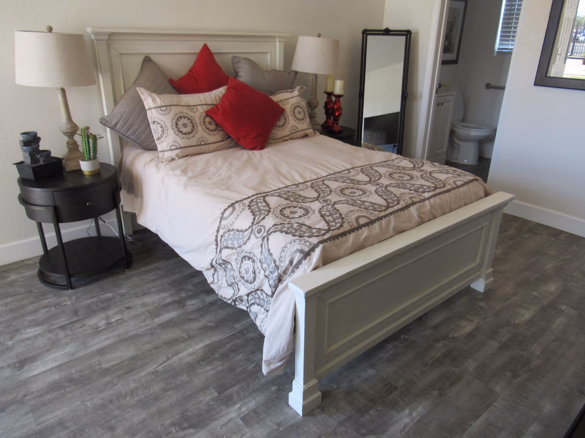 Well-lit bedroom with a white bed frame, patterned bedding and red accent pillows, a bedside table with a lamp, and an open door showing a bathroom.