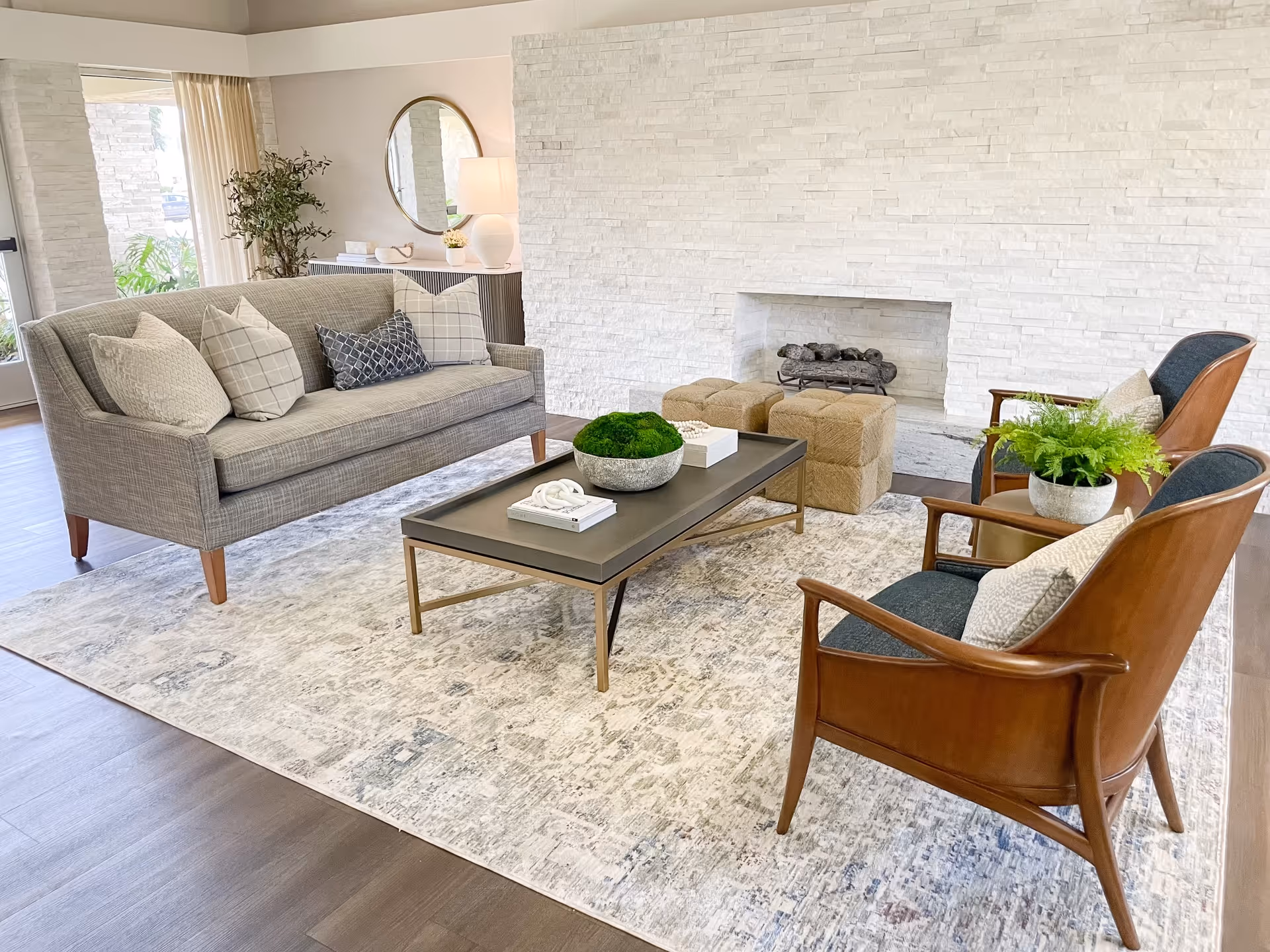 A modern living room with a gray sofa adorned with patterned pillows, two wooden armchairs with dark cushions and light pillows, a rectangular coffee table with decorative items, two beige ottomans, a white textured stone fireplace, a large area rug, and a console table with a round mirror and a lamp in the background.