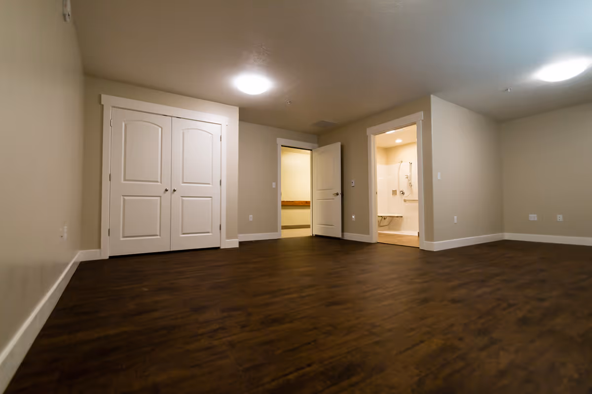 Empty room with dark wood flooring, beige walls, two closed white closet doors, an open door leading to a hallway, and another open door revealing a bathroom with a shower and grab bars.