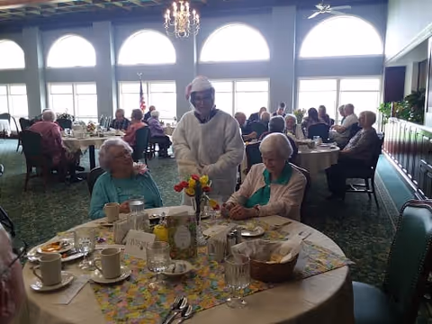 A group of elderly people seated at round dining tables in a bright room with large arched windows. One person wearing a festive hat is standing and interacting with two seated women at the foreground table, which is set with cups, plates, and a vase of flowers. Other tables with people are visible in the background.