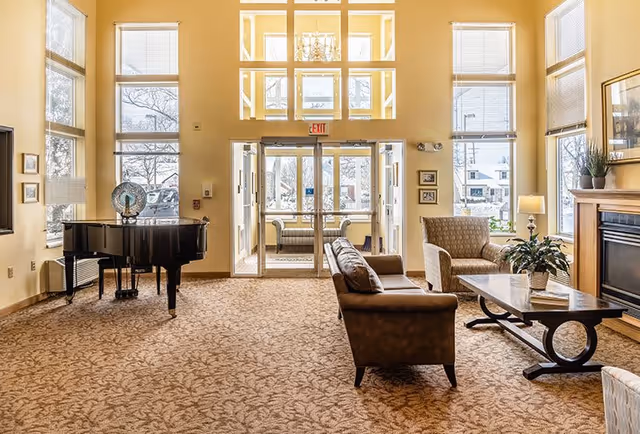 A spacious living room area with large windows allowing natural light to fill the space. The room features a grand piano on the left, a brown leather sofa and an armchair around a coffee table with a plant and books. There is a fireplace on the right side and glass double doors leading to another seating area with a couch. The walls are painted a warm beige color and the carpet has a patterned design.