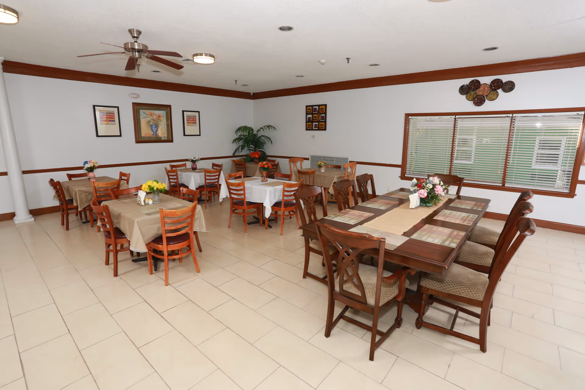 A dining room with several wooden tables and chairs arranged neatly. Some tables have beige tablecloths while others have white. Each table is decorated with a small flower arrangement. The room has tiled floors, white walls with brown trim, framed artwork, a ceiling fan, and a large window with blinds.