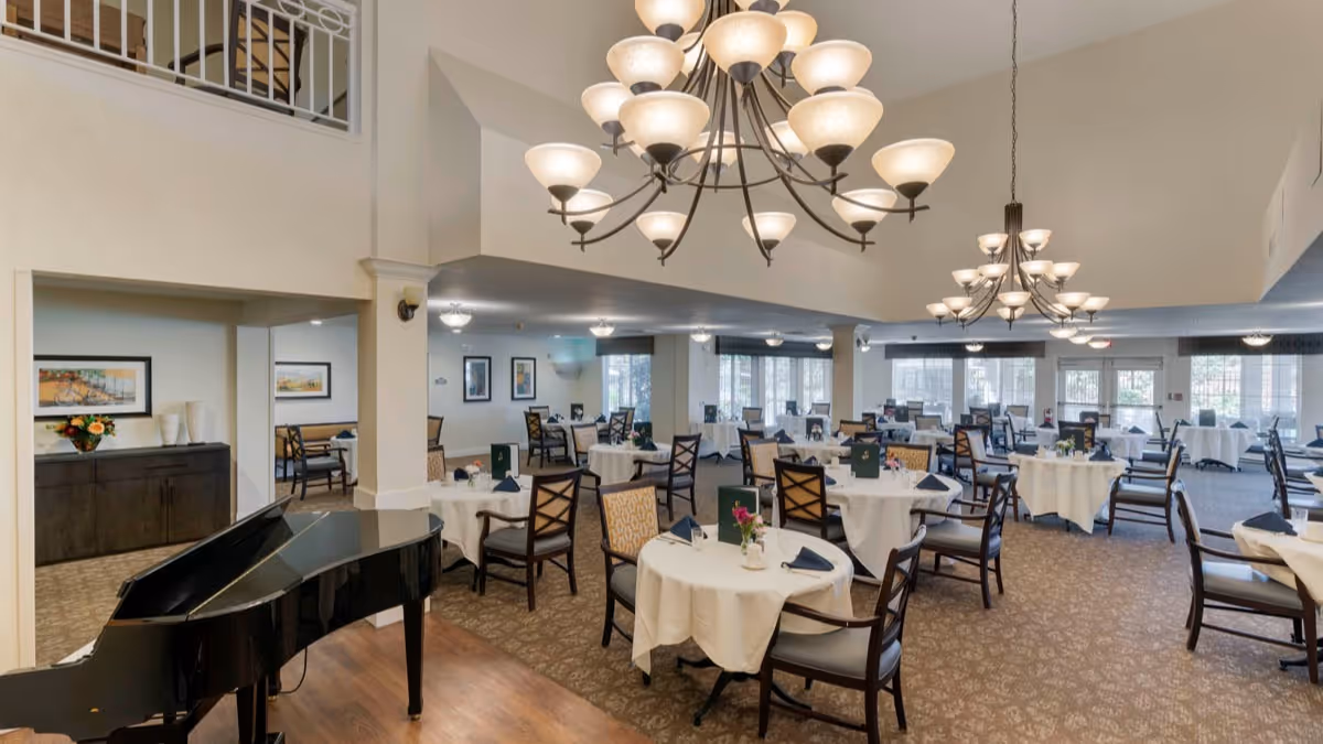 A spacious dining room with multiple round tables covered with white tablecloths, each set with napkins and small flower arrangements. There are wooden chairs around the tables, and large chandeliers hang from the ceiling. A black grand piano is positioned in the foreground on the left side. The room has large windows allowing natural light to enter and framed artwork on the walls.