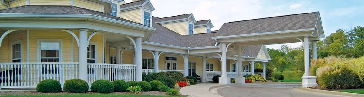 Exterior view of a yellow senior living facility building with white trim, a covered entrance supported by white columns, and well-maintained landscaping including bushes and grass under a partly cloudy sky.