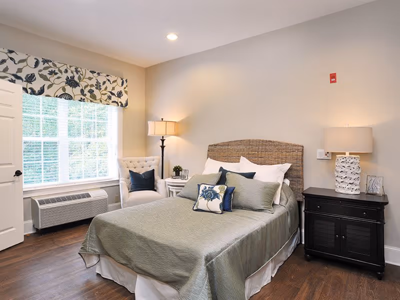 A cozy bedroom with a bed covered in a green bedspread and multiple pillows, including decorative ones with floral patterns. There is a wicker headboard, a white cushioned armchair with a dark pillow, a window with white blinds and a floral valance, a floor lamp, and a black nightstand with a textured white lamp and small decor items. The room has wooden flooring and beige walls.