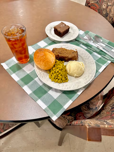 A meal served on a white plate with meatloaf, mashed potatoes, green peas, and a bread roll on a green and white checkered placemat. Next to the plate is a glass of iced tea and a small plate with a chocolate brownie. Silverware is wrapped in a green napkin on the side. The setting is on a round table with floral upholstered chairs.