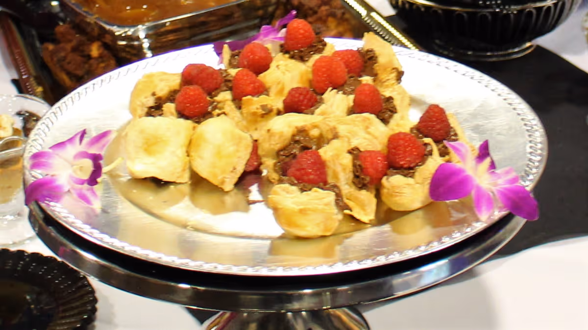 A silver platter with small puff pastry appetizers topped with chocolate spread and fresh raspberries, garnished with two purple orchid flowers on the sides.