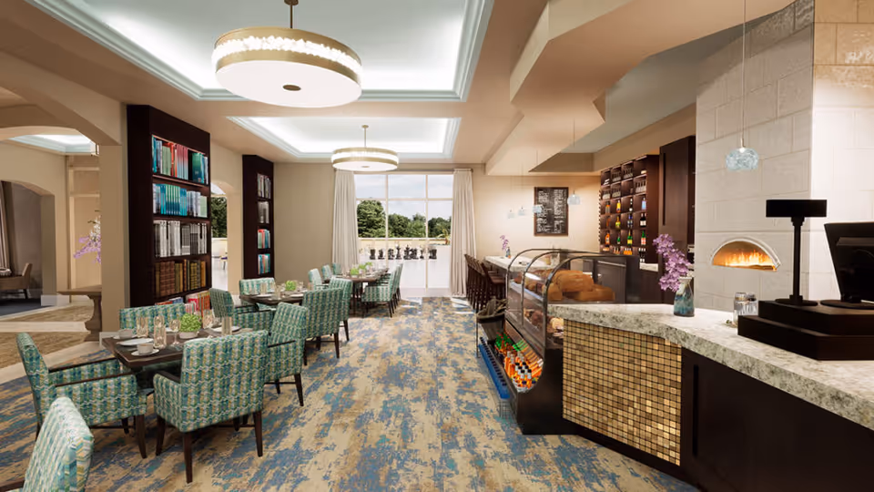 Bright dining and café area with patterned upholstered chairs and tables, a service counter with glass display case, and a decorative oven/fireplace.
