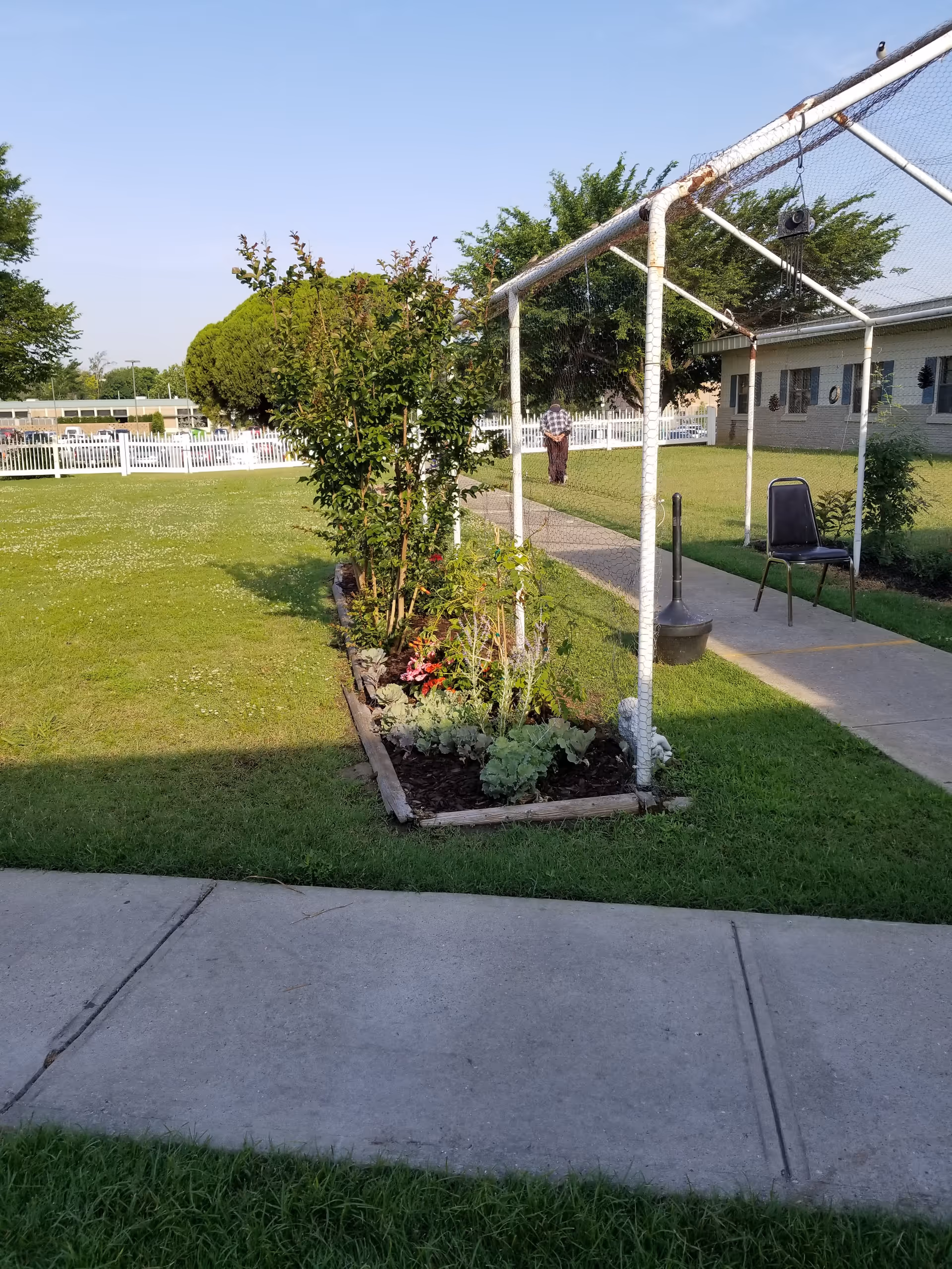 Outdoor lawn and garden area at a senior care facility with a trellis, paved walkway, a chair, and a person walking near a building.