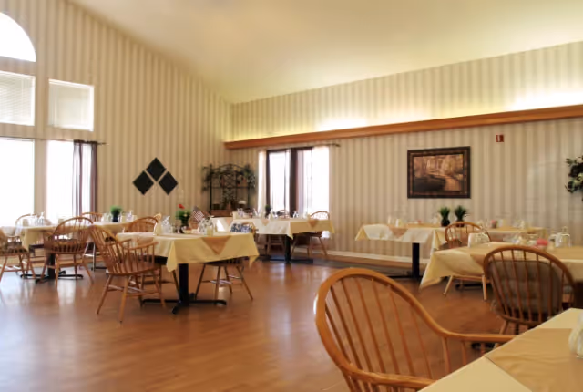 A dining room with several tables covered in beige tablecloths, each set with glasses and small flower arrangements. Wooden chairs surround the tables. The room has large windows with curtains, striped wallpaper, and a framed picture on the wall.