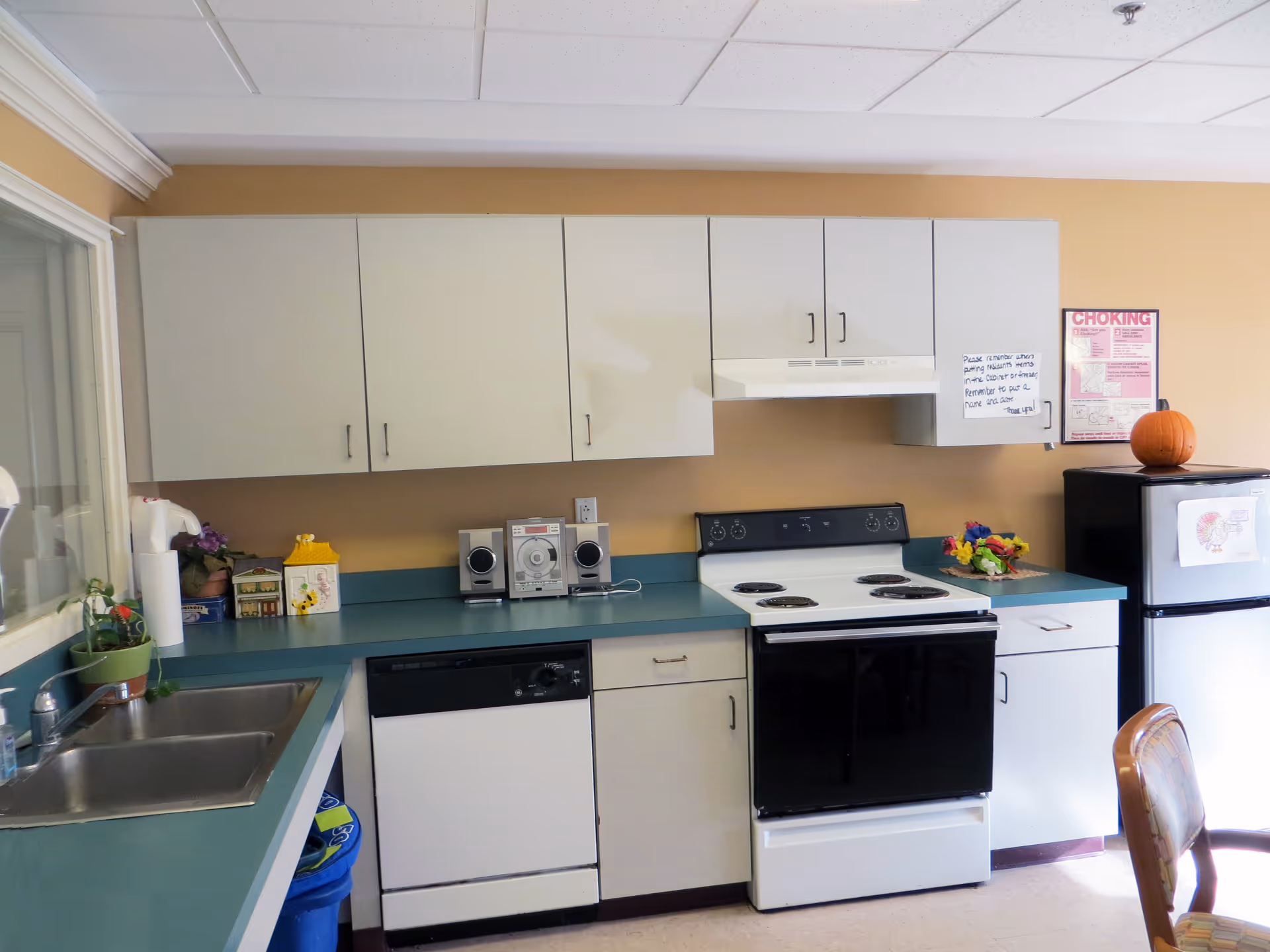 A kitchen area in a senior living facility with white cabinets, a green countertop, a white electric stove with an oven, a dishwasher, a refrigerator with a pumpkin on top, a sink, and various small items including a stereo and a colorful decoration. There is a chair partially visible on the right and a window on the left.