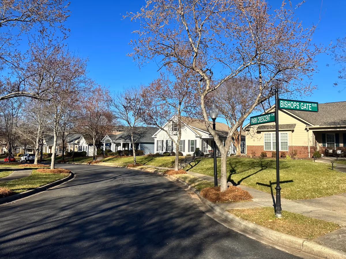 Sunny neighborhood street with single-story cottage-style homes, bare trees, and a street sign reading "Bishops Gate" and "Park Crescent."