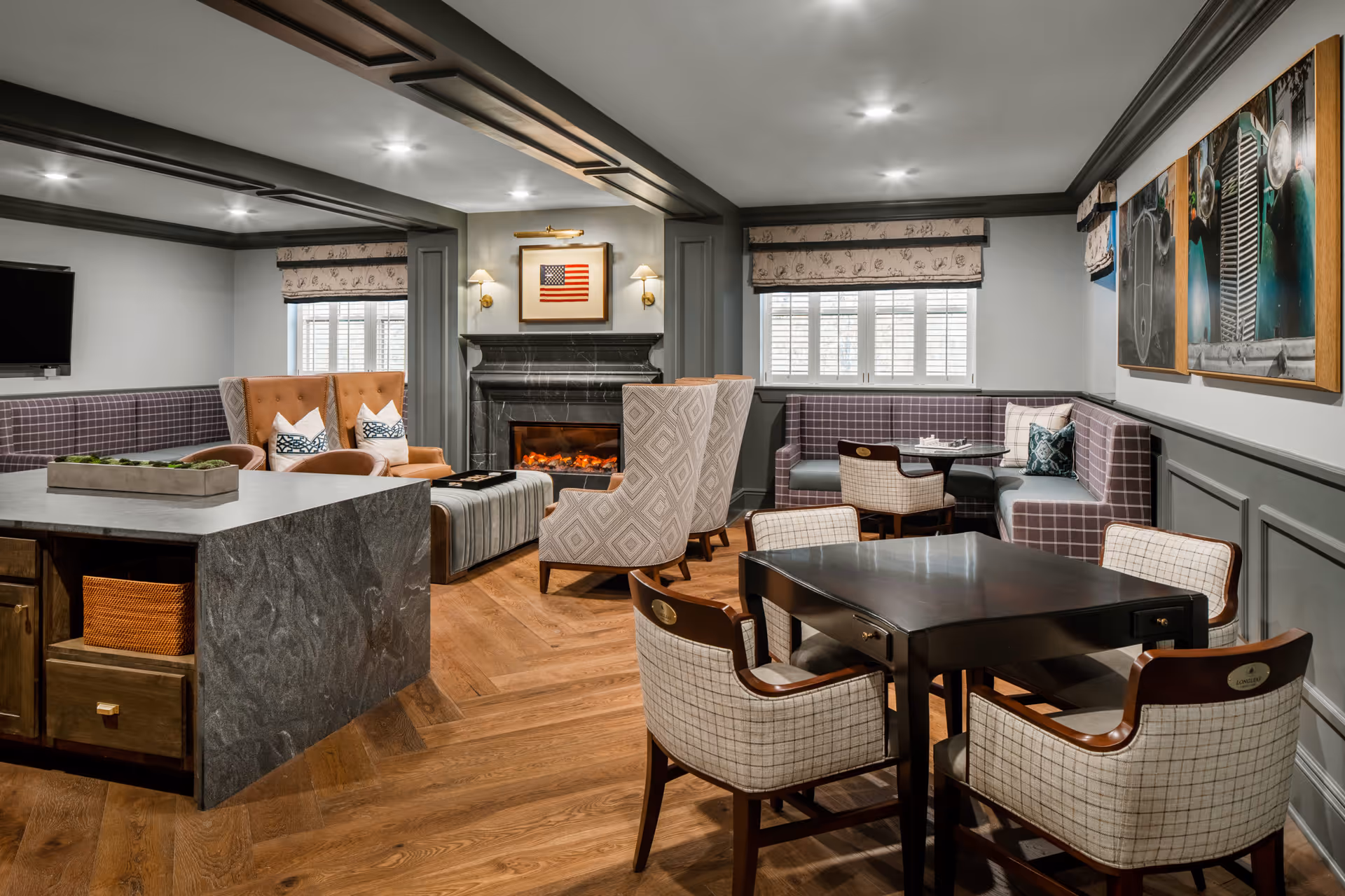 A cozy and well-lit common area featuring a fireplace with an American flag artwork above it, comfortable upholstered armchairs, a built-in bench with plaid cushions, a dark wooden table with four cushioned chairs, and a kitchen island with storage baskets underneath. The room has wooden flooring, gray wainscoting, and windows with patterned Roman shades.